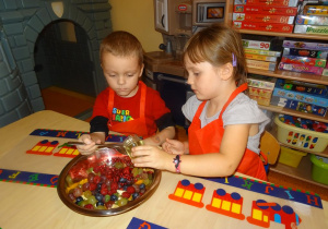 Dwoje dzieci siedzi przy stole. Dziewczynka trzyma w ręku słoik z miodem z którego chłopiec łyżką przekłada miód do miski z pokrojonymi owocami.